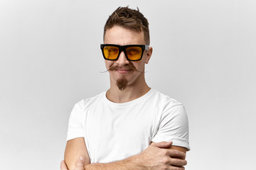 Cheerful attractive young unshaven male with stylish hairdo, goatee beard and handlebar mustache being in good mood, crossing arms on his chest and smiling confidently, wearing trendy spectacles - obrazy, fototapety, plakaty