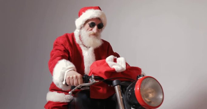 Excited santa claus with red bag full of christmas gifts getting ready to drive off on big black bike, concept of modern way of holiday celebration