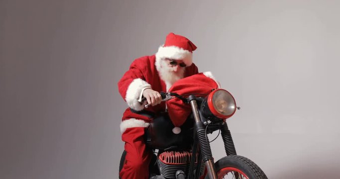 Santa claus in stylish round sunglasses gets on big black bike, isolated indoors