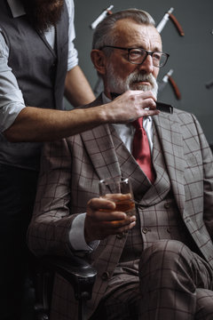 Barber making male haircut. Hairdresser cutting hair of his rich client, dressed in expensive stylish man s suit of clothes and holding a glass of alcoholic beverage