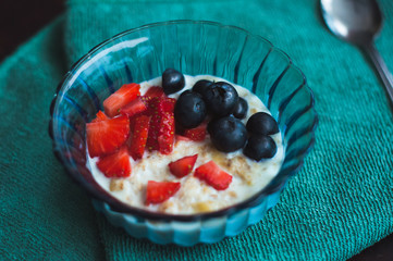 High angle view on cooked oat with forest fruits. Strawberries and blueberries in a blue glass bowl on a green napkin.