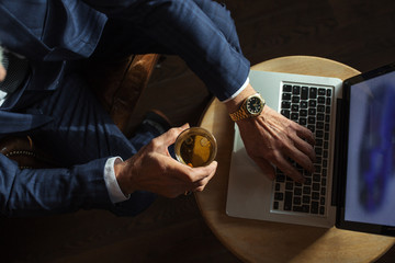 Old man s hands in stylish dark blue suit working on laptop while relaxing at restaurant while carrying glass of wisky, top view