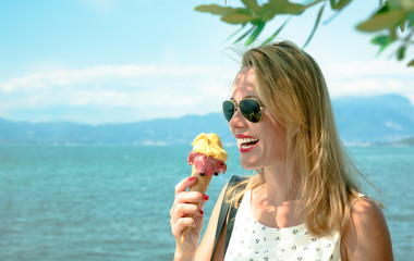 junge hübsche  blonde Frau mit rotem Hut am Meer See bei Sonnenschein und blauem Himmel macht Selfies mit Handy, ist ein Eis in der Waffel Waffeleis und lächelt