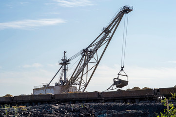 A huge excavator overloads in the dumps rock from the unloaded train from the mine against a clear blue sky. Concept: mining and environmental problems