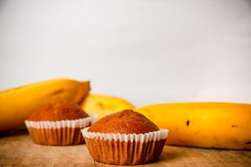 Closeup photo of the banana bread muffin in white paper cup and the bananas on wooden background. The homemade easy bake oven cake.