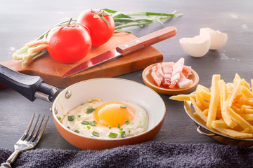 Fried egg in a pan, bacon, french fries tomatoes and green onions are cooked for breakfast on a wooden gray table. Close-up