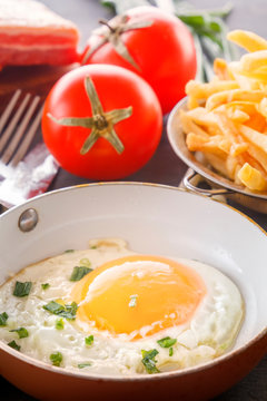 Fried egg in a pan, bacon, french fries tomatoes and green onions are cooked for breakfast on a wooden gray table. Close-up