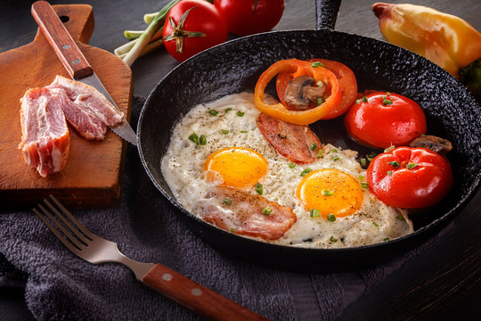 Fried eggs with bacon and tomatoes on an old cast iron pan and cutlery on a gray table. Close-up
