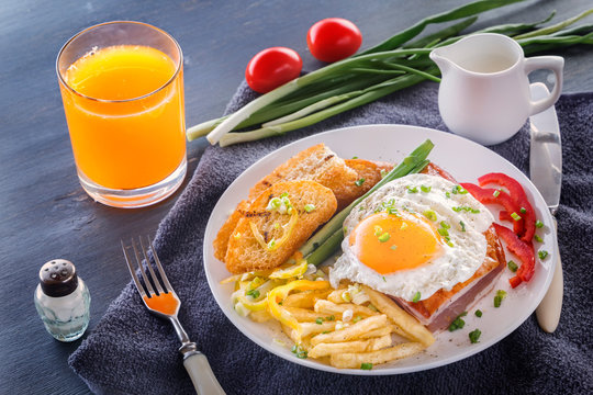 Fried egg with bacon in a white plate with fried pieces of bread, greens, tomatoes, french fries and a glass of fresh juice on a gray wooden table. Close-up