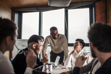 deadline, technology and people concept - creative team of multiracial men, sitting round the desk in diverse casual and formal clothes talking over new project in office meeting room