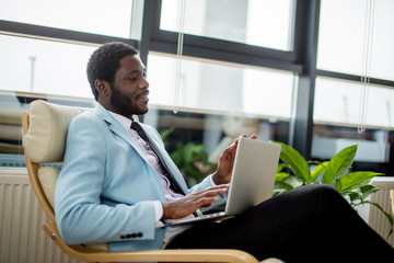 Successful African bearded entrepreneur in sky blue coat and tie serfing web sites with attentive and concentrated look, searching useful information