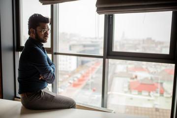 Confident, thinking business man of indian origin. Pensive person wearing a formal dark shirt poses against big window with cityscape on background