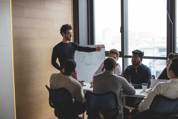 Fototapeta premium Young caucasian sales manager presenting new project to his multiethnic colleagues, reporting about work result with flipchart.