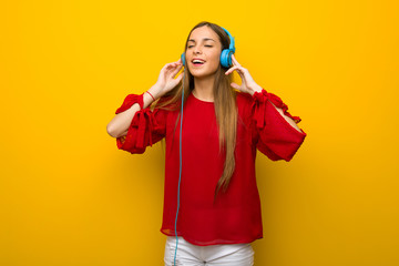 Young girl with red dress over yellow wall listening to music with headphones