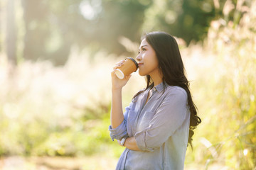 Young woman with cup of coffee on hand in the meadow at the morning time.