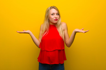 Young girl with red dress over yellow wall making unimportant gesture while lifting the shoulders