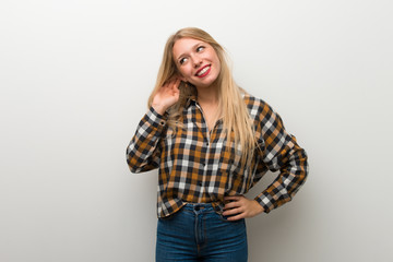 Blonde young girl over white wall listening to something by putting hand on the ear