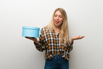 Blonde young girl over white wall holding gift box in hands