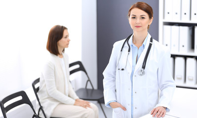 Doctor woman at work. Portrait of female physician cheerful smiling while standing near reception desk at clinic or emergency hospital. Patient woman sitting at the background. Medicine concept