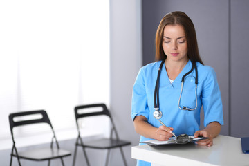 Fototapeta na wymiar Doctor woman at work. Portrait of female physician filling up medical form while standing near reception desk at clinic or emergency hospital. Medicine and healthcare concept