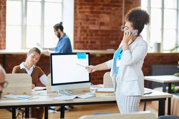Obraz premium Young confident office manager talking to client by smartphone while standing by computer and pointing at its screen