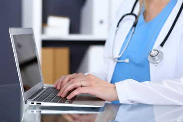 Doctor typing on laptop computer while sitting at the glass desk in hospital office. Physician at work. Medicine and healthcare concept