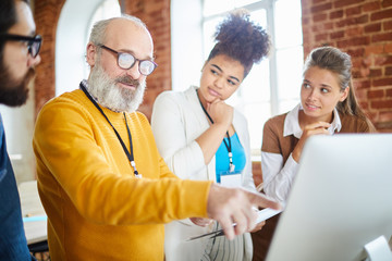 Confident mature man in eyeglasses pointing at computer screen during discussion of ideas for new business project with young subordinates