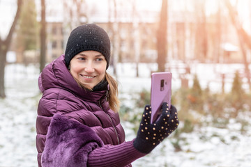 Young happy woman in warm purple dawn jacket smiling while making selfie with smartphone during walk in winter city park. Pretty girl enjoys winter vacations and travel