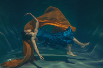 cute sporty female swims underwater as a free diver in a blue evening dress with orange fabric alone - Powered by Adobe