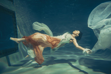 elegant slender girl swims underwater like a free diver in a white evening dress with beautiful...