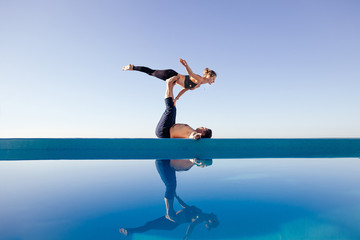 Acro yoga concept. Pair yoga. Couple of young sporty people practicing yoga lesson with partner, man and woman in yogi exercise, arm balance pose, working out by pool, above beach, against blue sky