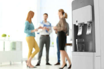 Modern water cooler with glass and blurred office employees on background. Space for text