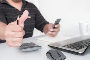 Male businessman's right hand Hold pencil and showing thumbs up and left hand pressing mobile phone. Calculator, mouse, computer, laptop placed on a white desk.