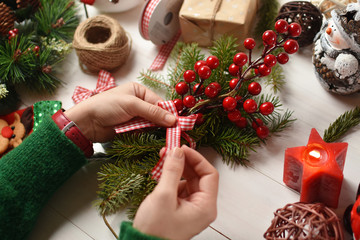 Female hands decorating fir branch with holly and ribbon bow at the desk surrounded with Christmas...