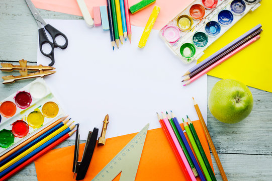 a cup with colored pencils, watercolor paints, a notebook, a ruler, a stapler, scissors on a blue wooden background, top-down view 