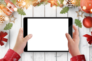 Child in Christmas sweater holding black tablet with empty screen, white wooden table with Christmas decorations in background
