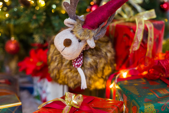Toy deer between presents and christmas tree on the background. Red and green boxes with different patterns and golden tapes. Blur lights. Closeup