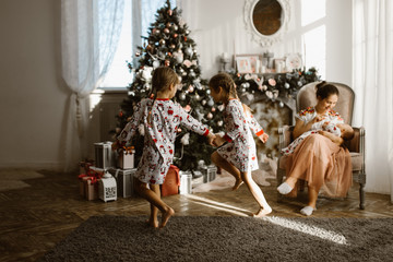 Two little sisters in pajamas having fun New Year's tree with gifts in the light cozy room and their mother sits in the armchair with little baby next to the fireplace