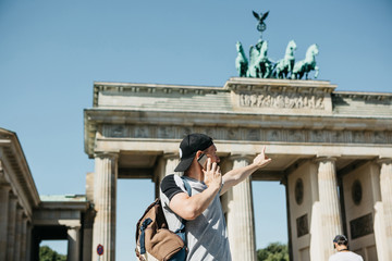 A tourist or a guy with a backpack is talking on a mobile phone and shows his hand on the Brandenburg Gate in Berlin in Germany and makes an appointment. © franz12