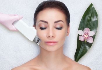 The doctor-cosmetologist makes the ultrasound cleaning procedure of the facial skin of a beautiful,...