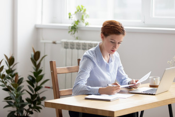 Concentrated serious businesswoman sitting at desk holding documents checking analyzing data, sales statistics doing paperwork. Confident company executive manager preparing report for conference