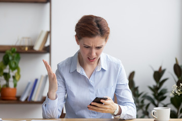 Stressed businesswoman sitting at working desk looking at mobile phone screen feels irritation...