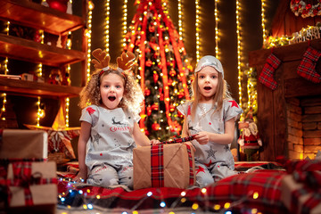 Happy little girls wearing Christmas pajamas playing, wondering with gift box by a fireplace in a cozy dark living room on Christmas eve. Celebrating Xmas at home.