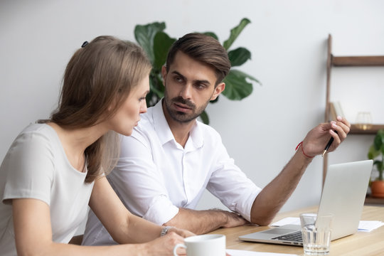 Serious millennial guy and girl company members workmates sitting together at office desk using computer discussing project having problems or different opinion and visions arguing during busy workday