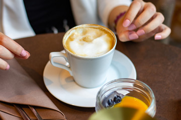 Brown table with fresh cappuccino coffe in a white cup, fruit dessert and young woman's hands- close up