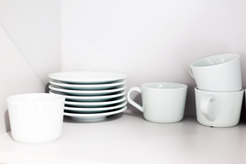 White ceramic coffee cups and saucers on shelves. Shelves with kitchen utensils.