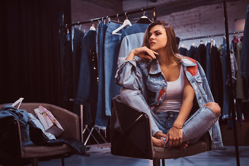 A stylish girl wearing distressed jeans and coat covering his shoulder sitting on a chair in the fitting room of a clothing store.