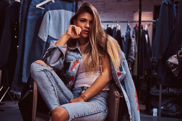 Obraz premium A seductive girl wearing a distressed jeans and denim jacket looking at a camera while sitting on a chair in the fitting room of a clothing store.