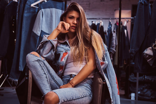 A seductive girl wearing a distressed jeans and denim jacket looking at a camera while sitting on a chair in the fitting room of a clothing store.