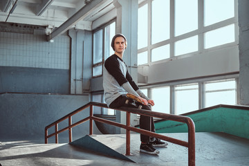 Young handsome man wearing shirt and hat holding his board while sitting on a grind rail in...